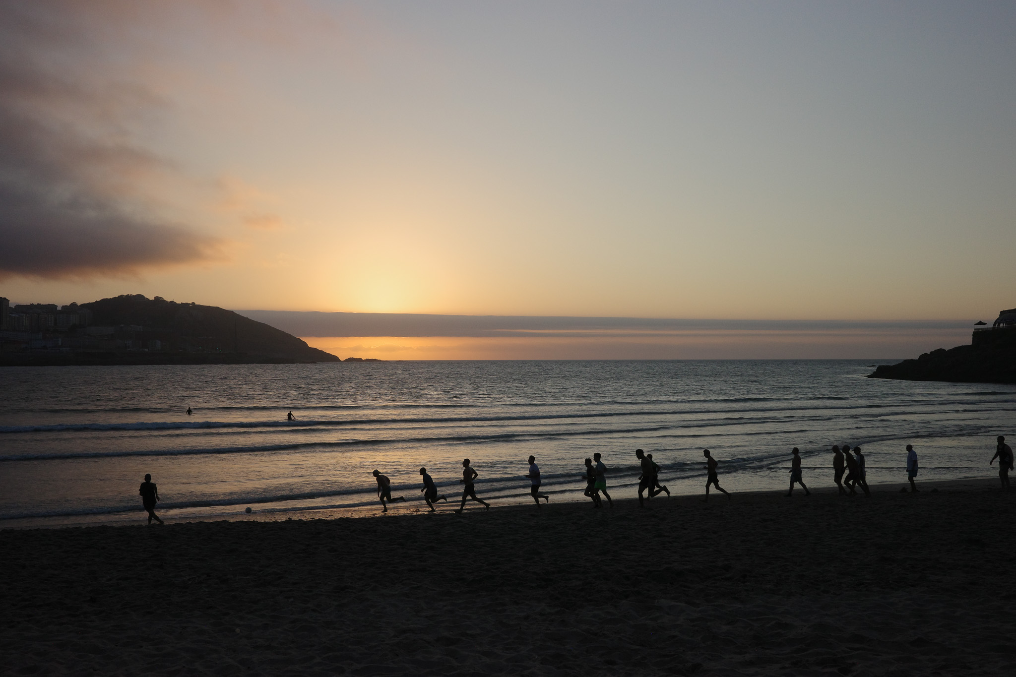 Un gruppo di bagnanti gioca a calcio lungo la spiaggia mentre il Sole si nasconde, con un cielo nuvoloso e una playlist di brani dedicati all’eclissi